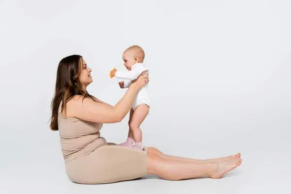 Side view of happy plus size mother holding baby with toy on grey background — Stock Photo