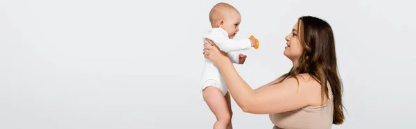 Side view of cheerful woman with overweight holding baby with toy isolated on grey, banner — Stock Photo