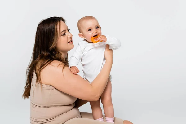 Morena mãe com sobrepeso segurando bebê com brinquedo isolado no cinza — Fotografia de Stock