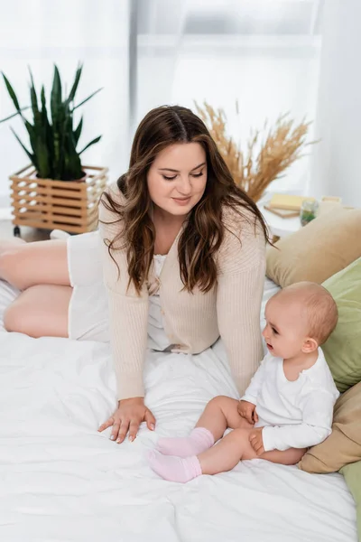 Young body positive mother looking at cheerful baby on bed — Stock Photo