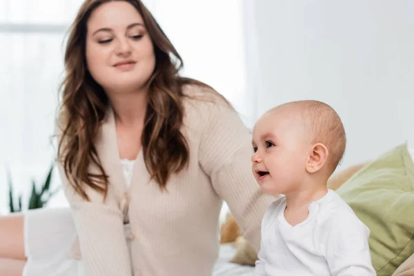 Cheerful baby girl sitting on bed near blurred mom at home — Stock Photo