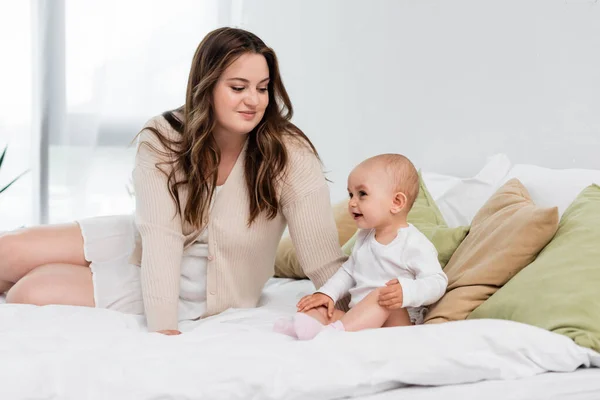 Счастливая мать plus size смотрит на улыбающегося ребенка на кровати в спальне — стоковое фото