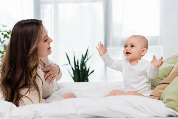 Cheerful baby daughter sitting near brunette mother on bed at home — Stock Photo