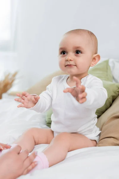 Baby girl sitting on bed near blurred hands of mother — Stock Photo