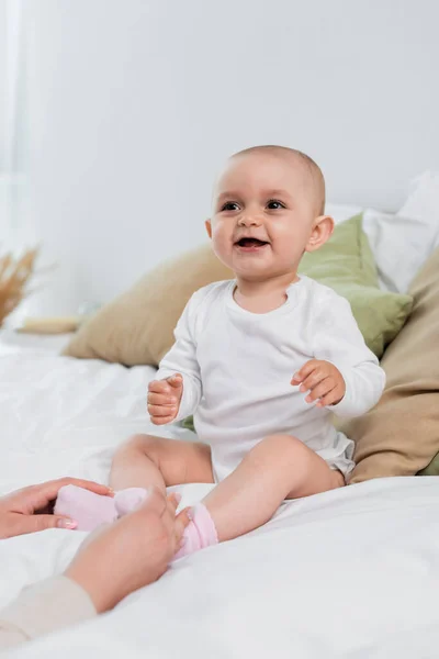 Joyeux bébé fille assis sur le lit près des mains de maman — Photo de stock