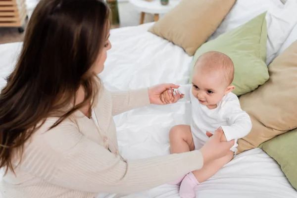 Vista de ángulo alto de la madre cogida de la mano de la hija bebé en la cama - foto de stock