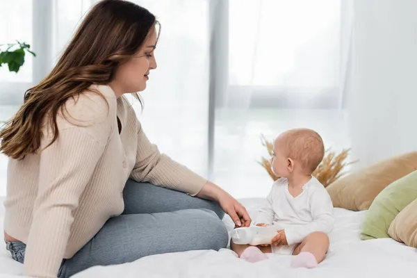 Pretty pus size woman sitting near baby daughter with bottle of milk on bed — Stock Photo