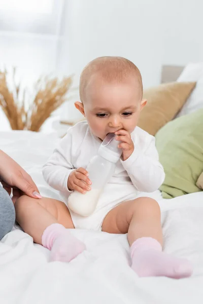 Bébé fille tenant le biberon avec du lait près de la mère au lit — Photo de stock