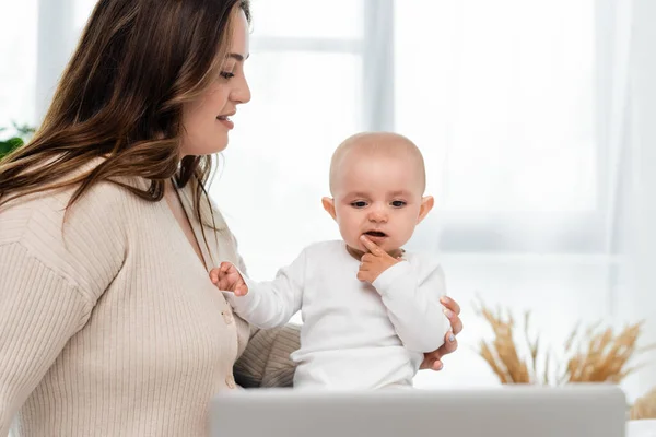 Smiling plus size mother looking at baby daughter near blurred laptop at home — Stock Photo