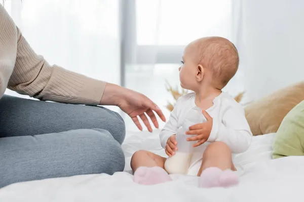 Young woman sitting near baby with bottle of milk on bed — Stock Photo