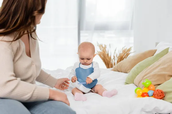 Giovane madre positiva del corpo seduta vicino al bambino con piatto e cucchiaio sul letto — Foto stock