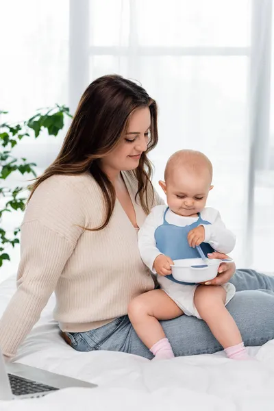 Souriant plus taille femme assise près de bébé fille avec plaque et cuillère et ordinateur portable sur le lit — Photo de stock