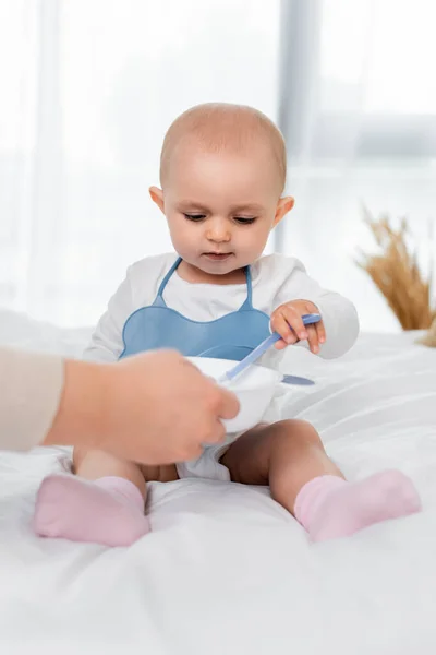 Woman holding plate with baby food near daughter with spoon on bed — Stock Photo
