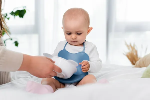 Woman holding plate near baby daughter on bed at home — Stock Photo