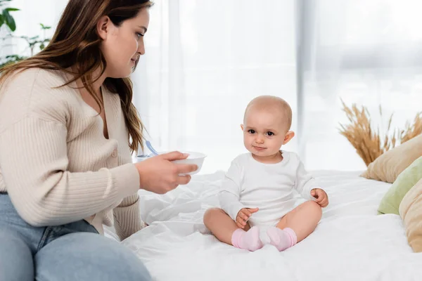 Plus-Size-Mutter hält Teller neben Baby-Tochter auf Bett — Stockfoto