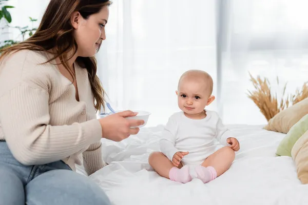 Plus-Size-Mutter hält Teller neben Baby-Tochter auf Bett — Stockfoto