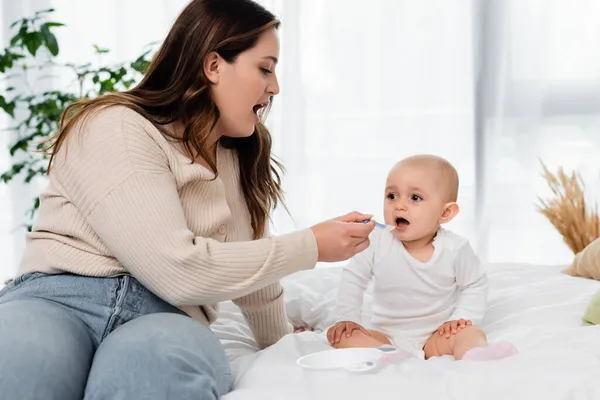 Plus size mother open mouth while feeding baby on bed — Stock Photo