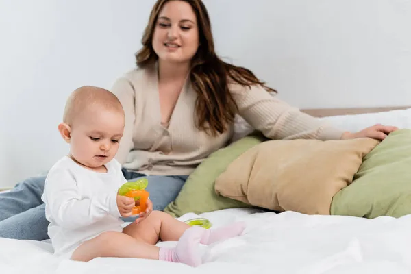 Bambino ragazza giocare con giocattoli vicino sfocato plus size madre sul letto — Foto stock