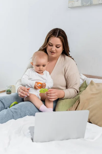 Cuerpo joven mujer positiva sosteniendo juguete cerca del bebé y el ordenador portátil en la cama - foto de stock