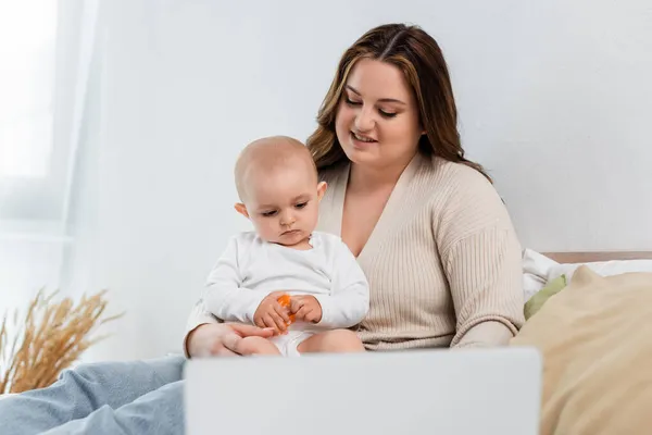 Sonriente madre de tamaño grande mirando al niño con juguete cerca de la computadora portátil borrosa en la cama - foto de stock