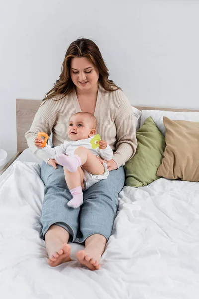 Positivo más tamaño madre mirando al bebé con juguetes en la cama - foto de stock