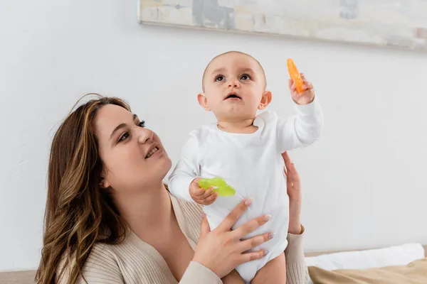 Mujer joven con sobrepeso sosteniendo bebé niña con juguetes en casa - foto de stock