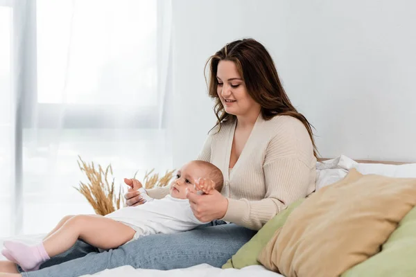 Positive plus size woman touching hands of baby daughter on bed — Stock Photo