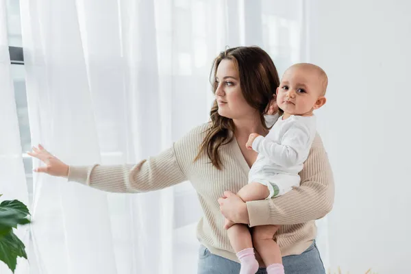 Mulher bonita com sobrepeso segurando bebê perto de cortinas em casa — Fotografia de Stock