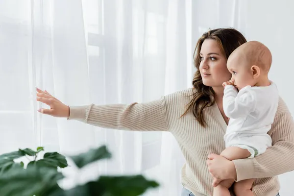 Young body positive mother holding child near curtains at home — Stock Photo