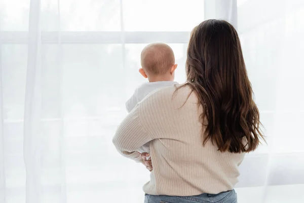 Vista posteriore della donna plus size che tiene il bambino vicino alla finestra a casa — Foto stock