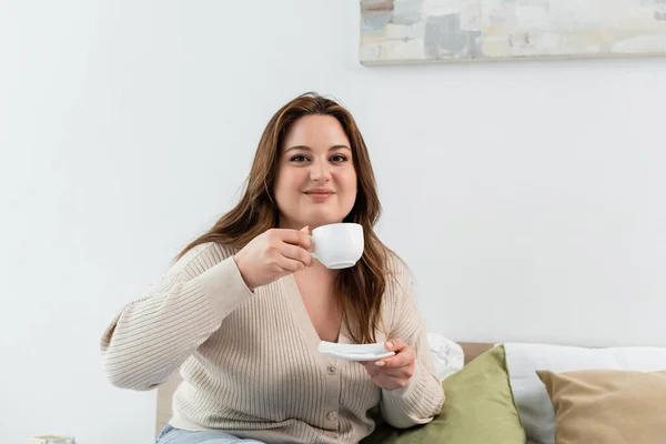 Pleased body positive woman holding cup and saucer on bed at home — Stock Photo