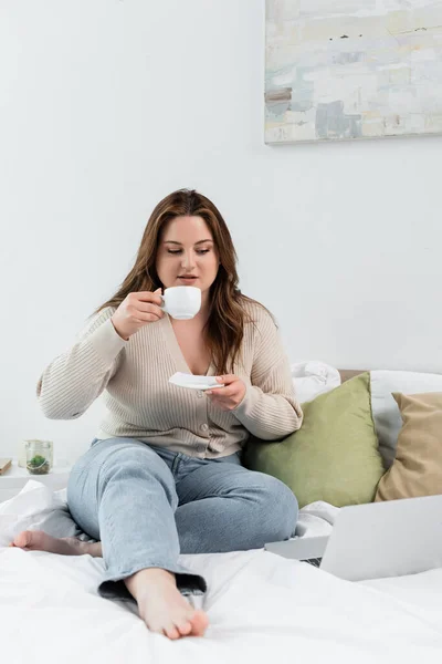 Молодая женщина с избыточным весом держа чашку кофе возле ноутбука на кровати — стоковое фото