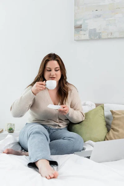 Mujer joven con sobrepeso sosteniendo taza de café cerca de la computadora portátil en la cama - foto de stock