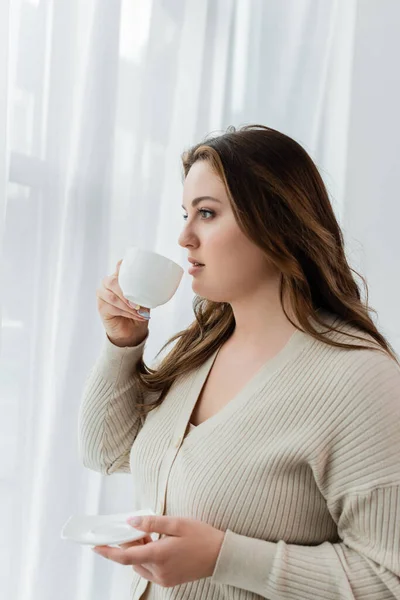 Joven más tamaño mujer sosteniendo la taza de café cerca de las cortinas en casa - foto de stock