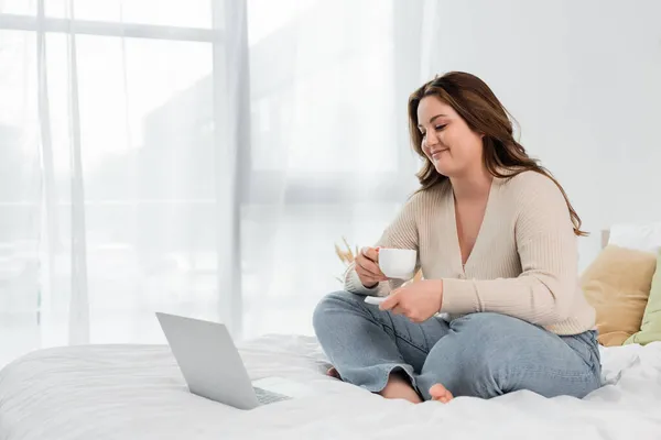 Smiling body positive freelancer holding cup while using laptop on bed — Stock Photo