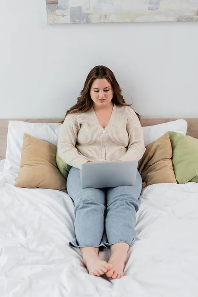 Jolie femme positive du corps travaillant sur un ordinateur portable sur le lit à la maison — Photo de stock