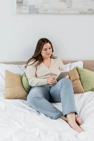 Bruna donna con libro di lettura in sovrappeso sul letto — Foto stock