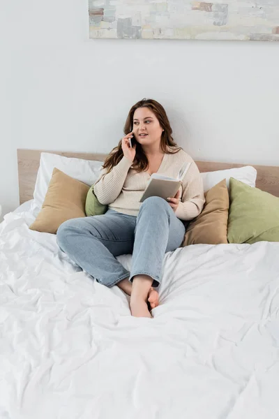 Sorridente plus size donna parlando sul telefono cellulare e tenendo libro sul letto — Foto stock