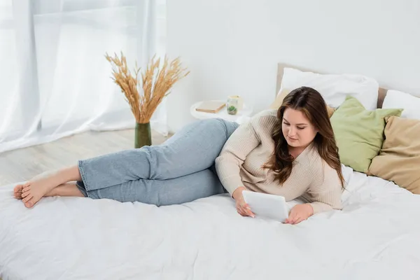 Barefoot plus size woman using digital tablet on bed — Stock Photo