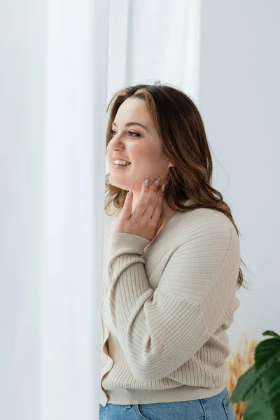 Positivo plus tamanho mulher olhando para cortinas em casa — Fotografia de Stock