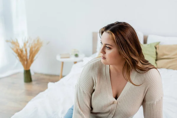 Mujer joven con sobrepeso mirando hacia otro lado en la cama borrosa en casa - foto de stock