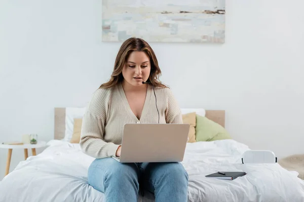 Freelancer com excesso de peso usando laptop e fone de ouvido perto notebook na cama — Fotografia de Stock