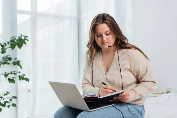 Junge Freiberuflerin mit Übergewicht schreibt auf Notebook, während sie Laptop und Headset im Schlafzimmer benutzt — Stockfoto