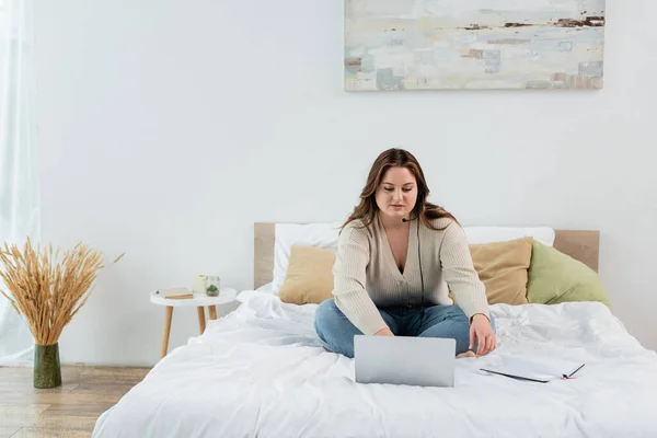 Plus size donna in auricolare utilizzando laptop vicino notebook sul letto — Foto stock