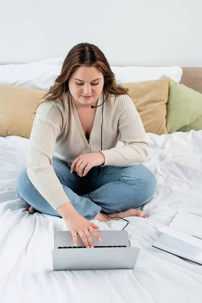 Cuerpo teletrabajo positivo en auriculares usando portátil cerca de portátil en la cama - foto de stock