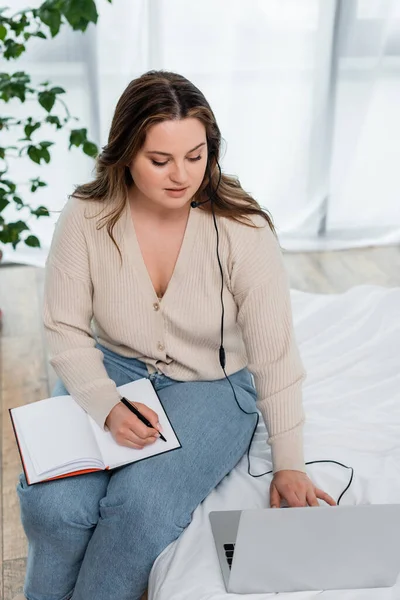 Pretty body positive freelancer in headset writing on notebook and using laptop on bed — Stock Photo