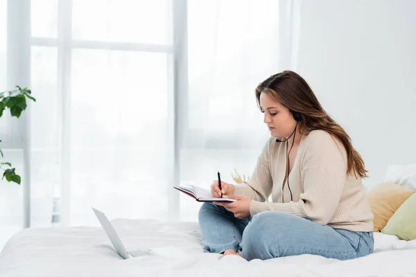 Young body positive freelancer in headset writing on notebook near laptop in bedroom — Stock Photo