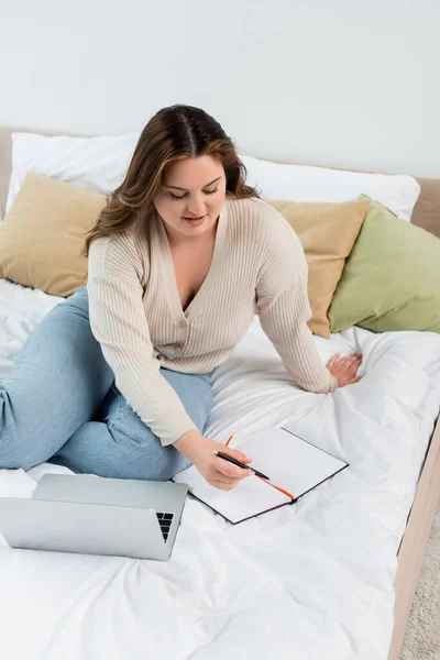 Lächelnde Freiberuflerin hält Stift neben Notizbuch und Laptop auf dem Bett — Stockfoto