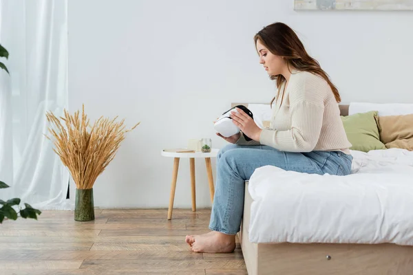 Vue latérale du jeune corps positif femme exploitation vr casque sur le lit à la maison — Photo de stock