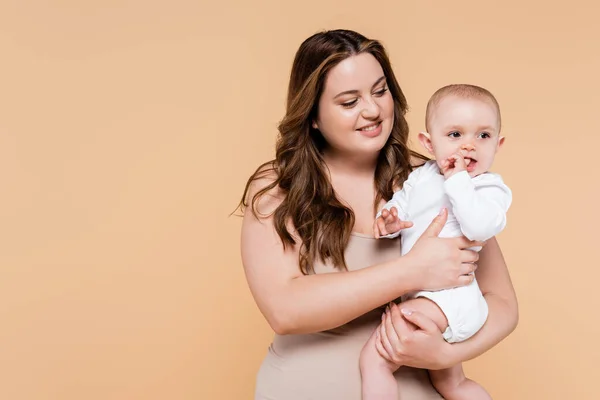 Sonriente cuerpo positivo madre sosteniendo al bebé con el dedo cerca de la boca aislado en beige - foto de stock